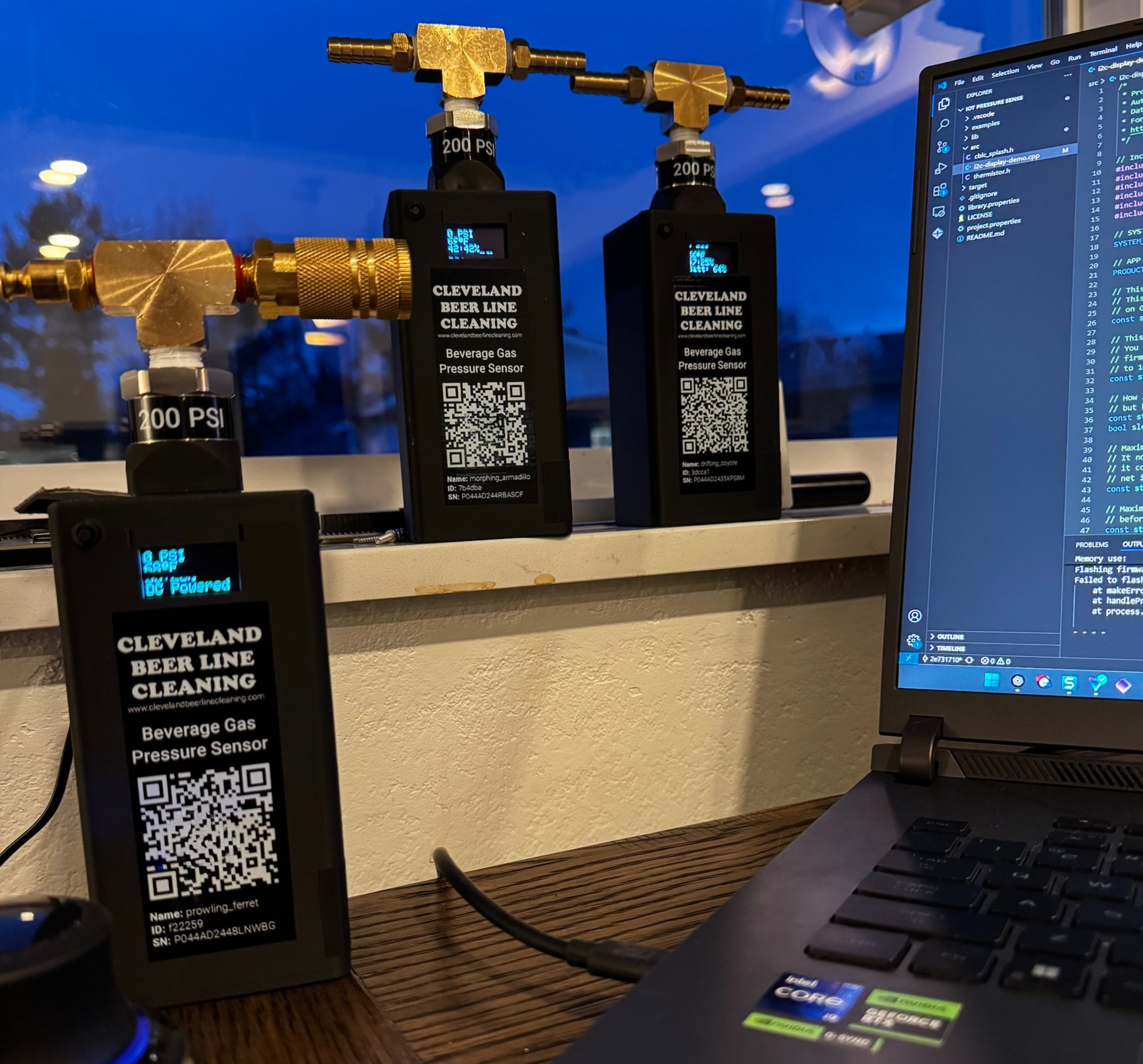 Three early Barkeep beverage gas pressure sensors on a windowsill showing live PSI readings, next to a laptop running the sensor firmware in a code editor.